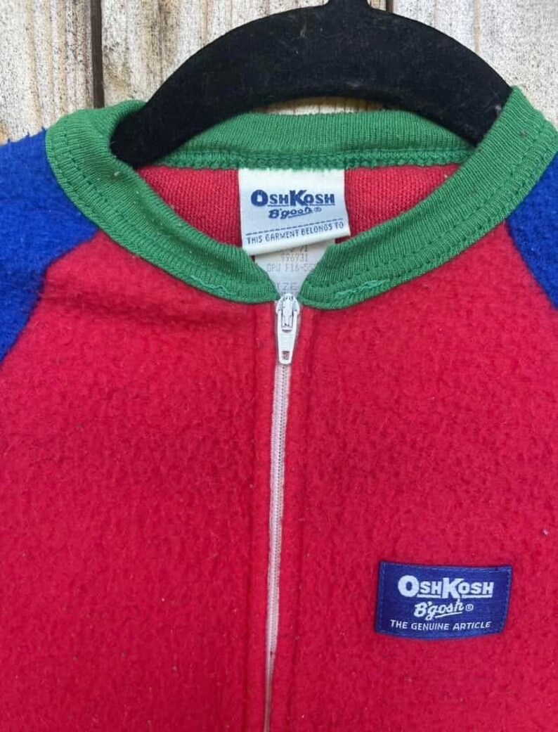 Puede incluir: Un mono vintage OshKosh B'gosh de una pieza en rojo, azul y verde. El mono tiene una cremallera completa, un cuello verde y mangas azules. La etiqueta OshKosh B'gosh es visible en el pecho. La prenda est&aacute; colgada en una percha negra.
