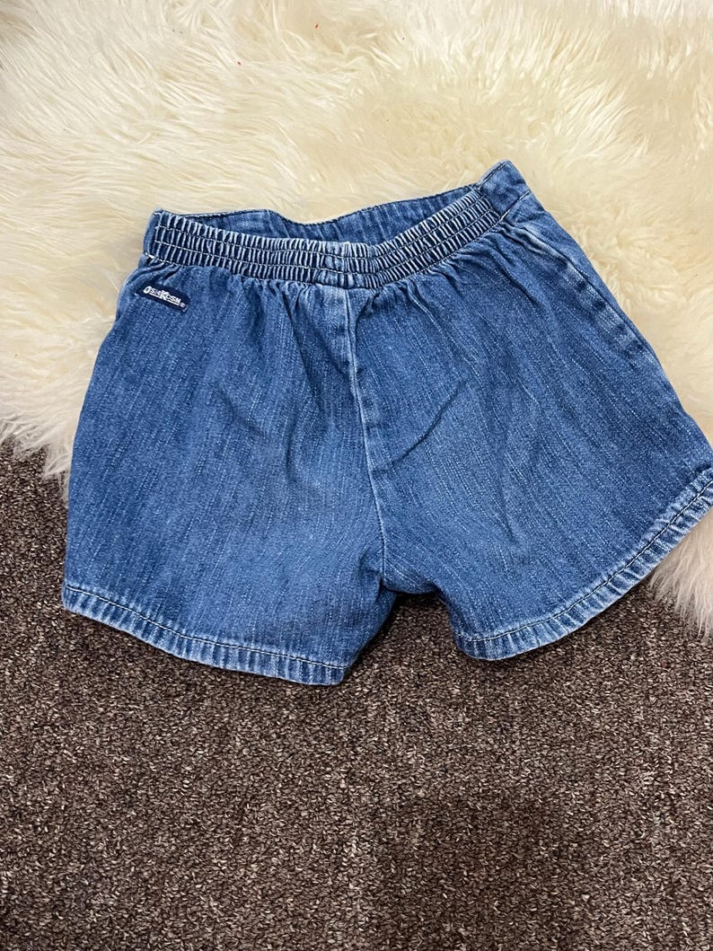 May include: Blue denim shorts with an elastic waistband. The shorts have a classic five-pocket design and are made of a durable denim fabric. The shorts are likely for a child.