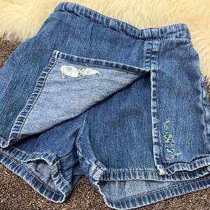 May include: Blue denim skort with floral embroidery. The skort features a wrap-style front with a decorative embroidered floral design on the edge. The shorts have a classic denim look with a comfortable fit.