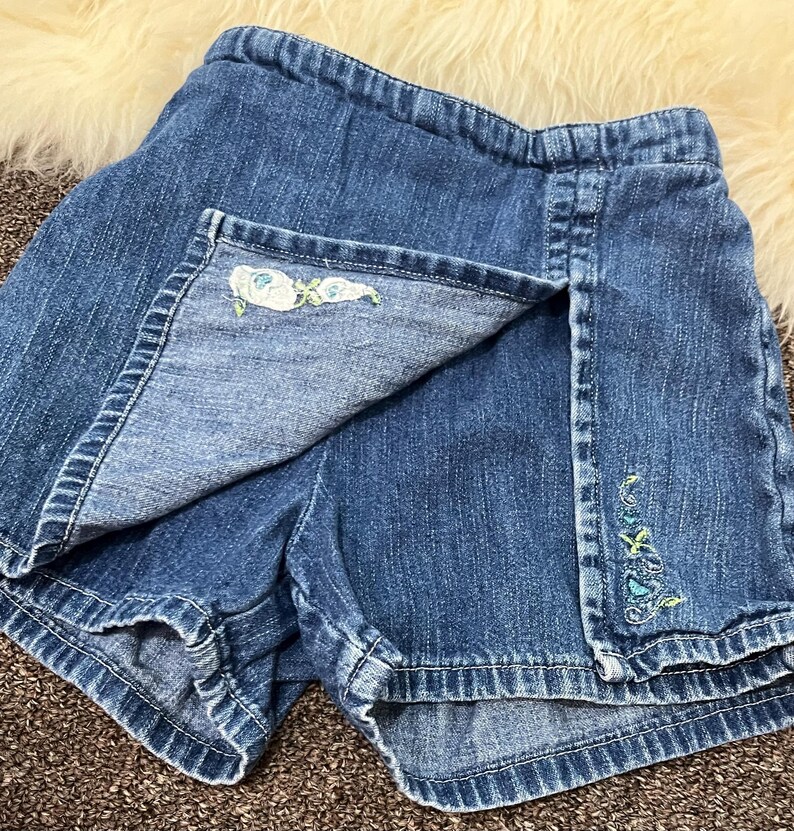 May include: Blue denim skort with floral embroidery. The skort features a wrap-style front with a decorative embroidered floral design on the edge. The shorts have a classic denim look with a comfortable fit.