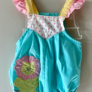 May include: A baby romper in turquoise blue with a lion applique. The romper has pink and yellow accents on the shoulders and ruffled edges. The bodice features a white panel with pink polka dots.