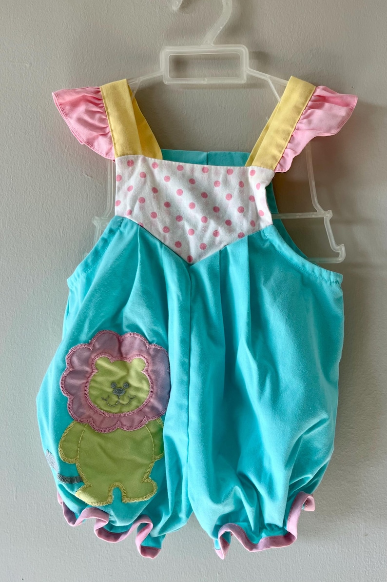 May include: A baby romper in turquoise blue with a lion applique. The romper has pink and yellow accents on the shoulders and ruffled edges. The bodice features a white panel with pink polka dots.