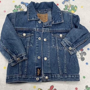 May include: A blue denim jacket with a button-up front, two chest pockets, and two side pockets. The jacket has a collared neckline and long sleeves with buttoned cuffs. The brand label "OshKosh B'Gosh" is visible on the jacket.