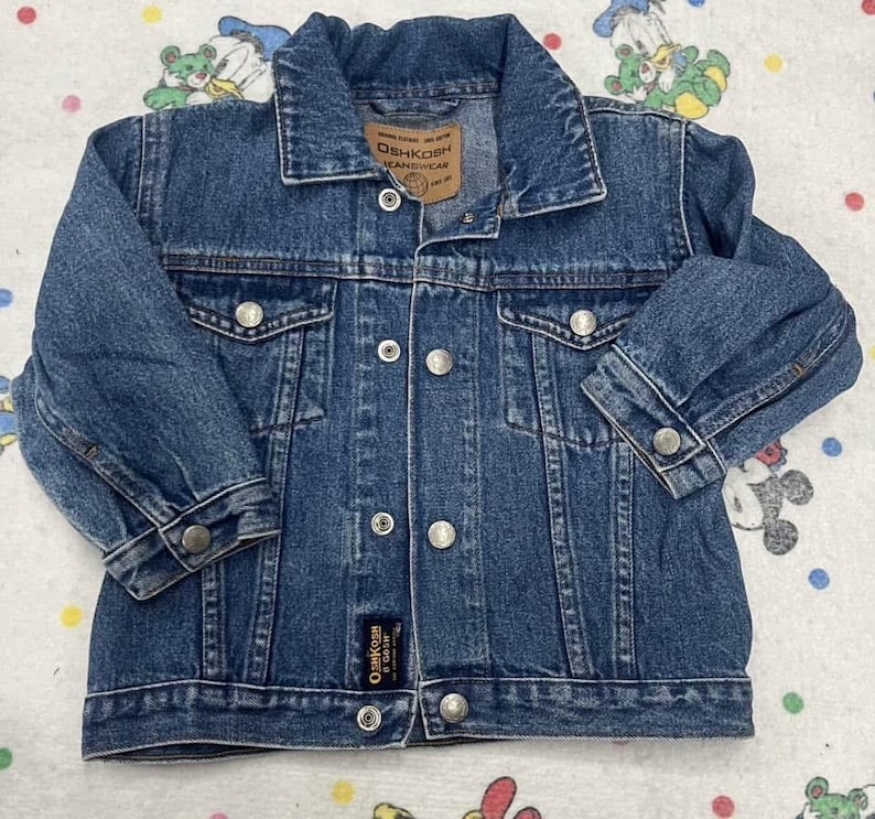 May include: A blue denim jacket with a button-up front, two chest pockets, and two side pockets. The jacket has a collared neckline and long sleeves with buttoned cuffs. The brand label "OshKosh B'Gosh" is visible on the jacket.