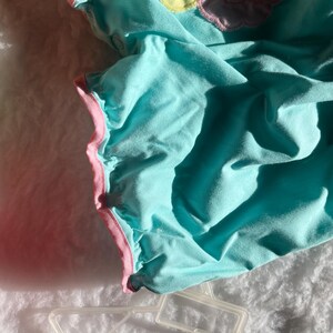 May include: A turquoise baby romper with pink trim and a ruffled edge. The romper has a soft, textured appearance and is lying on a white, fluffy surface. The garment is partially visible, with shadows adding depth.