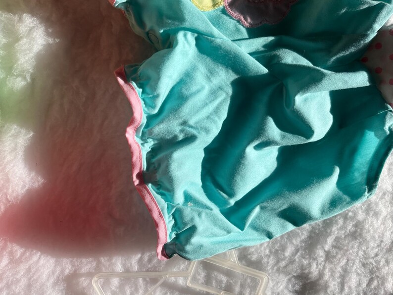 May include: A turquoise baby romper with pink trim and a ruffled edge. The romper has a soft, textured appearance and is lying on a white, fluffy surface. The garment is partially visible, with shadows adding depth.