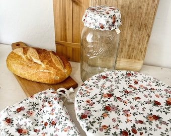Puede incluir: Un juego de tres cubiertas de alimentos reutilizables con un patrón floral. Las cubiertas están hechas de tela y tienen un cierre de cordón. Las cubiertas se muestran sobre una superficie blanca con una hogaza de pan y un frasco de vidrio.