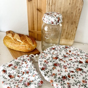 May include: A set of three reusable food covers with a floral pattern. The covers are made of fabric and have a drawstring closure. The covers are shown on a white surface with a loaf of bread and a glass jar.