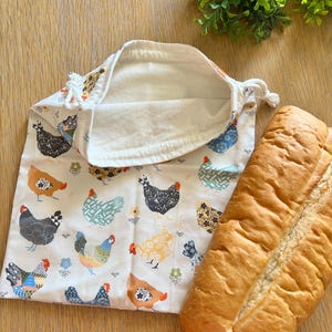 Puede incluir: Una bolsa de tela blanca reutilizable para productos con cierre de cord&oacute;n. La bolsa presenta un patr&oacute;n colorido de pollos con dise&ntilde;os geom&eacute;tricos. Una hogaza de pan blanco est&aacute; sentada junto a la bolsa.