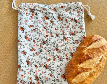 Puede incluir: Una bolsa de algodón blanco con cordón de ajuste con un estampado floral en tonos rosa, rojo y verde. La bolsa está llena de una hogaza de pan blanco.