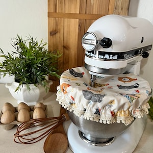 May include: White KitchenAid stand mixer with a stainless steel bowl. The bowl has a fabric cover with a chicken print and a white lace trim. There are eggs, a wooden spoon, and a whisk in the foreground.