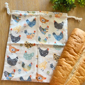 May include: A white cotton reusable produce bag with a drawstring closure. The bag features a repeating pattern of colorful chickens with floral accents. A loaf of white bread is sitting next to the bag.