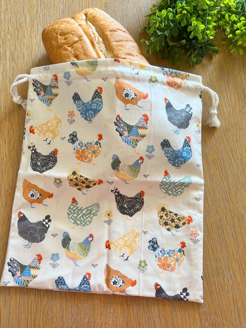 Puede incluir: Una bolsa de tela blanca reutilizable para productos con cierre de cord&oacute;n. La bolsa presenta un patr&oacute;n repetitivo de pollos coloridos con dise&ntilde;os florales y geom&eacute;tricos.