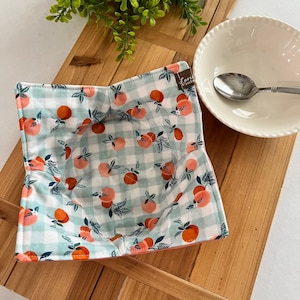 Peut inclure: Un protège-bol en tissu gingham bleu clair et blanc avec un imprimé pêche et vert. Le protège-bol est plié et posé sur une surface en bois.