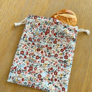 May include: A white reusable produce bag with a floral pattern in shades of red, pink, and yellow. The bag is made of cotton and has a drawstring closure. A loaf of bread is partially inside the bag.