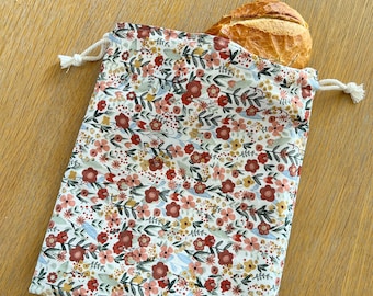Puede incluir: Una bolsa de tela reutilizable blanca con un estampado floral en tonos rojos, rosas y amarillos. La bolsa está hecha de algodón y tiene un cierre de cordón. Una barra de pan está parcialmente dentro de la bolsa.