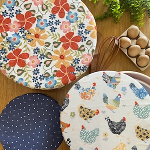 May include: Three reusable food wraps with different patterns. One wrap has a floral pattern with red, yellow, blue, and pink flowers on a white background. Another wrap has a navy blue background with white polka dots. The third wrap has a white background with a repeating pattern of colorful chickens.