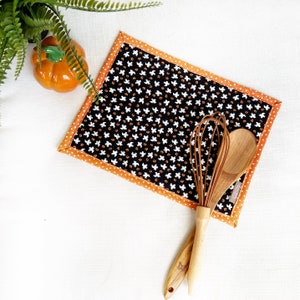 May include: A rectangular black and orange Halloween placemat with a white ghost pattern. A wooden whisk and spoon rest on the placemat. A small orange pumpkin and green plant are in the background.
