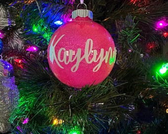 May include: Pink Christmas ornament with the name "Kaylynn" written in white cursive lettering.