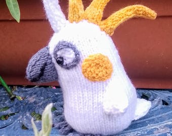 May include: A handmade knitted cockatoo bird plush toy. The bird is primarily white with yellow crest feathers, a gray beak, and gray feet. It has a round orange patch on its cheek and a black eye. The plush is sitting on a blue metal surface.