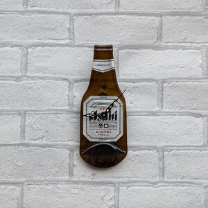 May include: A beer bottle-shaped wall clock with the "Asahi" logo and Japanese text. The clock has black hands and is mounted on a white brick wall. The bottle is brown with a white label.