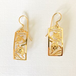 May include: Gold tone earrings with a cutout design of two sea turtles. The earrings are rectangular in shape and have a dangling hook.