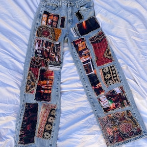 May include: Light blue denim jeans featuring a wide-leg silhouette and a high-waisted fit. The front of the pants is decorated with various rectangular fabric patches in dark, multi-colored patterns, all secured with visible black stitching.
