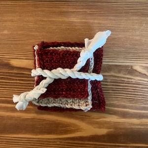 May include: A stack of square crocheted coasters in alternating deep red and beige colors, secured together with a thick, white, twisted cotton rope tied in a simple knot across the center of the fabric squares.