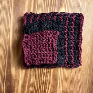 May include: A square, hand-crocheted washcloth featuring a textured, ribbed pattern in alternating stripes of deep burgundy and dark navy blue. A smaller, solid burgundy crocheted square is layered on top of the larger, striped piece.