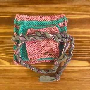 May include: A stack of three square knitted swatches in alternating pink and teal colours, featuring a speckled texture. A braided cord made of metallic purple and grey fabric strips is wrapped around the centre of the knitted squares.