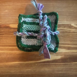 May include: A stack of square knitted coasters featuring horizontal stripes in shades of light green, dark green, and white. The coasters are bound together with a braided tie made of lavender and grey fabric strips knotted at the center.