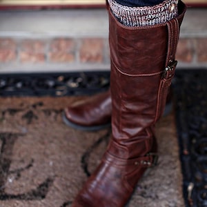 May include: Brown leather knee-high boots with a buckle detail. The boots are worn with a patterned sock.