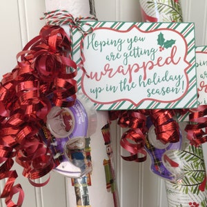 May include: A red and green striped gift tag with the text "Hoping you are getting wrapped up in the holiday season!"  The tag is tied to a roll of wrapping paper with red curling ribbon and a roll of tape.