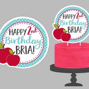 May include: A round cake topper with a blue and white polka dot background and red apples. The topper says "Happy 2nd Birthday BRIA!"