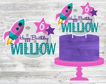 Puede incluir: Pastel morado con un soporte para pastel rosa. El pastel está decorado con un cohete, estrellas y una luna con el texto "Happy Birthday Willow".