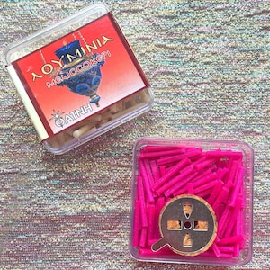 May include: A clear plastic container filled with pink incense sticks. The container also includes a metal incense burner with a hole in the center. The container has a label that reads "Aoyminia Megiccokeri  Datnh".