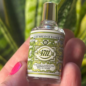 Pode incluir: Um pequeno frasco de vidro transparente de colónia com uma tampa prateada. O rótulo é verde e branco com o texto "ECHT KÖLNISCH WASSER ORIGINAL EAU DE COLOGNE N:4711. Eau de Cologne & Parfümerie-Fabrik "GLOCKENGASSE NO 4711" KOLN a. RH./COLOGNE"