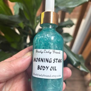 May include: A teal-colored body oil bottle with a gold cap and white dropper. The label reads "Morning Star Body Oil" and "Shady Lady Brand". The bottle is held in a hand, with green foliage in the background.
