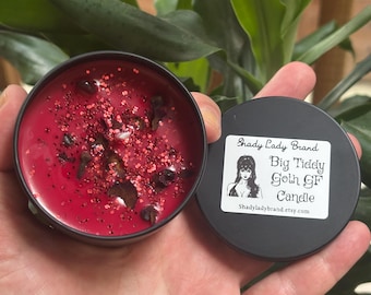 May include: A red candle in a black tin with glitter and dark stones. The lid features a graphic of a woman and the text "Big Tiddy Goth GF Candle" and "Shady Lady Brand".