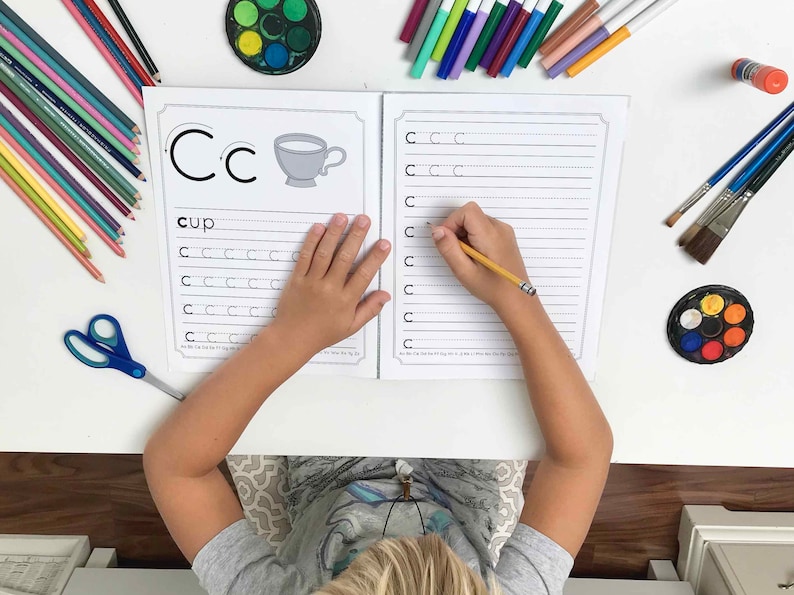 May include: A child is working on a handwriting worksheet for the letter 'C'. The worksheet has a picture of a cup and the word 'cup' written below it. The child is using a pencil to write the letter 'C' on the lines of the worksheet.