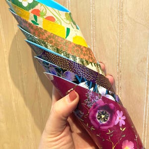 May include: A hand holds a stack of decorative paper cones with floral and fruit patterns. The cones feature designs with strawberries, lemons, and flowers in shades of red, yellow, purple, and blue. The cones are made of paper.