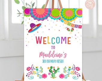 May include: A colorful welcome sign for a 3rd birthday fiesta. The sign features a sombrero, maracas, and colorful paper flowers. The text on the sign reads "Welcome to Madeleine's 3rd Birthday Fiesta!"