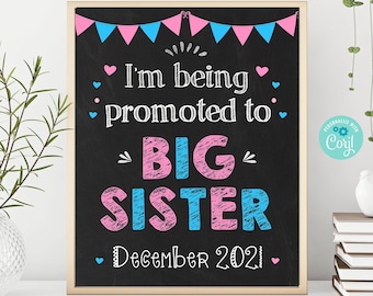 May include: A framed chalkboard sign with the text "I'm being promoted to BIG SISTER December 2021." The sign has pink and blue accents, including bunting and hearts, and is likely for a pregnancy announcement.