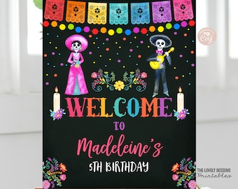 May include: Welcome to Madeleine's 5th Birthday sign with a black background, colorful flowers, and a skeleton couple in traditional Mexican Day of the Dead attire. The sign features a rainbow banner and colorful polka dots.