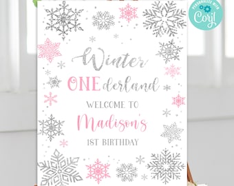 May include: A white sign with a silver glitter snowflake border and pink glitter text. The text reads "Winter One derland Welcome to Madison's 1st Birthday".