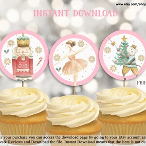 Puede incluir: Tres toppers redondos para cupcakes de 5 cm de diámetro con un Cascanueces, una bailarina y un ratón sosteniendo un árbol de Navidad. Los toppers están impresos en papel blanco con bordes rosas y detalles de copos de nieve dorados. El texto "IMPRIMIR 12 EN UNA PÁGINA" y "TAMAÑO 2" está impreso debajo de los toppers.