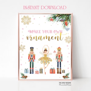 以下が含まれることがあります： ピンクの背景と金色の文字で「あなただけのオーナメントを作ろう」と書かれた印刷可能なサイン。サインには、くるみ割り人形、バレリーナ、オーナメントで飾られたクリスマスツリーが描かれています。