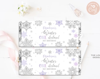 May include: Two rectangular party favor wrappers with a winter theme. The wrappers are white with silver and lavender snowflakes. Text reads "Madison's Winter ONE derland 1st Birthday".