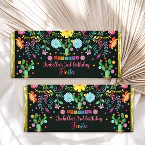 May include: Two chocolate bar wrappers with a black background, adorned with colorful floral and cactus designs. The text reads "Isabella's 3rd birthday Fiesta". Gold foil edges add a touch of elegance.