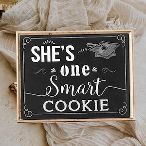 May include: A black chalkboard sign with a gold frame. The sign says "She's one smart cookie" with a graduation cap illustration.